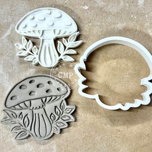 May include: Three white mushroom-shaped cookie cutters. One cutter is a complete mushroom design with a dotted cap and leaf accents. Another is a detailed outline of a mushroom. The third is a simple outline of a mushroom shape.
