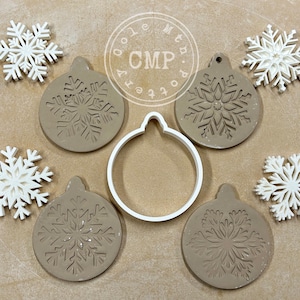 May include: A set of seven cookie cutters, including four round ornament shapes with snowflake designs, a round circle cutter, and two snowflake cutters. The cutters are made of white plastic and are on a brown surface.
