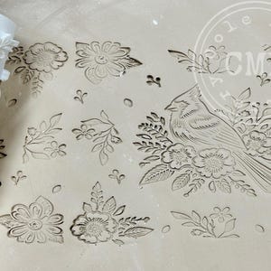 May include: A white roller with a floral and bird design. The roller is used to create a decorative pattern on a surface. The roller is next to a white surface with the same pattern imprinted on it.
