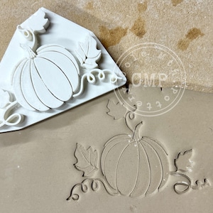 May include: A white, triangular-shaped pumpkin stamp with a detailed pumpkin design, leaves, and vines. The stamp is pressed into a clay surface, creating an embossed pumpkin image. The image is ideal for fall-themed crafts.