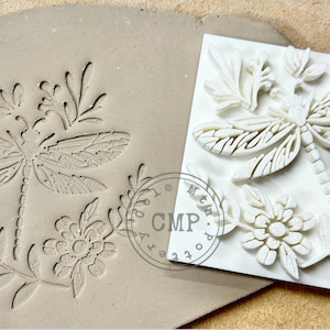 May include: A white rubber stamp with a detailed design of a dragonfly and floral elements. The stamp is on a light brown surface.