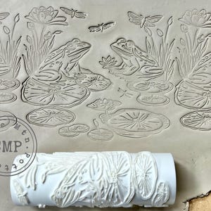 May include: A white rolling pin with a raised design of frogs, lily pads, and dragonflies. The rolling pin is being used to create an impression in a gray clay surface.