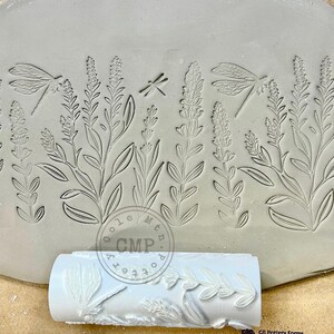 May include: A close-up of a clay tile with an embossed design of lavender flowers and dragonflies. The tile is being rolled with a rolling pin that has a similar design. The rolling pin is white and has the text "Cole Mtn. Pottery CMP" on the side.
