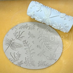 May include: A white rolling pin with a dragonfly and floral design. The rolling pin is used to create an imprint on a clay surface. The clay surface has a dragonfly and floral design.