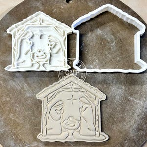 May include: Three white plastic cookie cutters in the shape of a nativity scene. Two are detailed with figures and a star, while one is a simple outline. The cutters are on a neutral-toned surface.