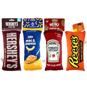 May include: Four plush dog toys shaped like food products. They include a Hershey's chocolate bar, Kraft Mac & Cheese, Heinz Tomato Ketchup, and Reese's Peanut Butter Cups. Each toy has a tag with the brand name and product details.