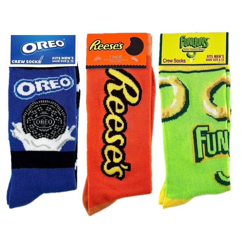 May include: Three pairs of crew socks featuring popular snack food brands. The socks are blue with an Oreo cookie design, orange with a Reese's Peanut Butter Cup design, and green with a Funyuns Onion Rings design. Each pair has the brand name and "Crew Socks" printed on the packaging.