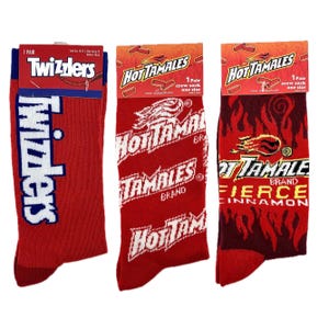 May include: Three pairs of crew socks featuring different brand logos. The first pair features a blue and white Twizzlers logo on a red background. The second pair features a white Hot Tamales logo on a red background. The third pair features a white Hot Tamales logo with a red and black flame design on a red background.