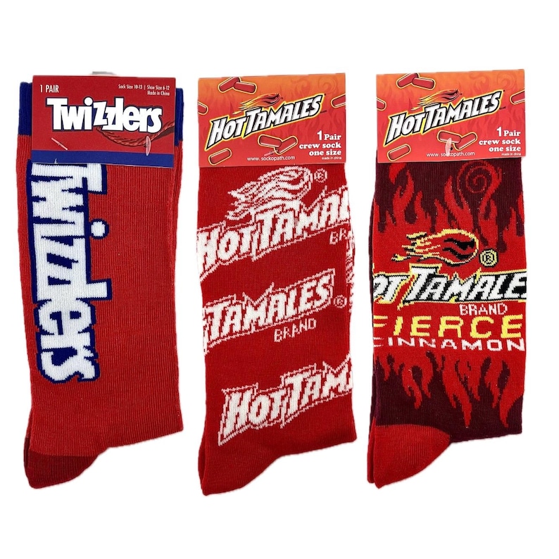 May include: Three pairs of crew socks featuring different brand logos. The first pair features a blue and white Twizzlers logo on a red background. The second pair features a white Hot Tamales logo on a red background. The third pair features a white Hot Tamales logo with a red and black flame design on a red background.