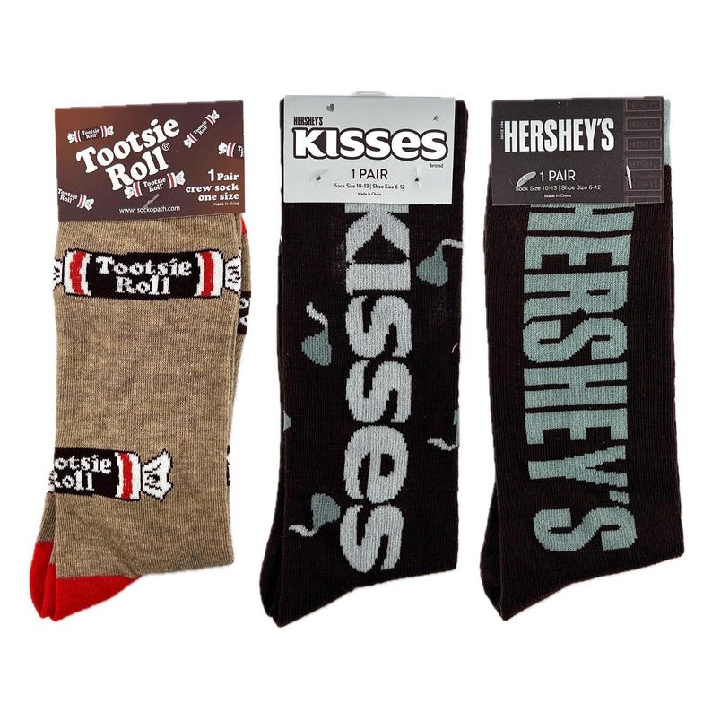 May include: Three pairs of crew socks featuring popular candy brands. The first pair is brown with the words "Tootsie Roll" printed in red and white. The second pair is black with the words "KISSES" printed in white. The third pair is black with the words "HERSHEY'S" printed in white.