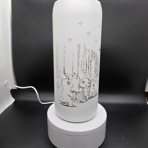 May include: White insulated tumbler with a silver lid. The tumbler features a black and white illustration of cartoon characters in a forest setting, with stars scattered across the upper portion. The tumbler is cylindrical and sits on a white rotating base.