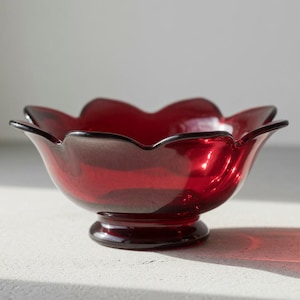 May include: A ruby red glass bowl with a scalloped rim and a pedestal base. The bowl's design resembles a flower, with each petal-like curve adding to its elegant shape. The bowl is made of transparent glass.