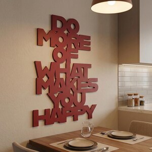 Könnte beinhalten: Rote Holz-Wanddekoration mit dem Schriftzug "DO MORE OF WHAT MAKES YOU HAPPY". Das Kunstwerk ist an einer beigefarbenen Wand über einem Holztisch angebracht. Eine Deckenleuchte beleuchtet den Essbereich.