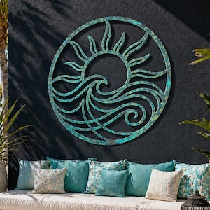 May include: A circular metal wall art piece featuring a stylized sun and ocean wave design. The sculpture has a weathered, oxidized teal and copper patina finish with visible texture, mounted on a dark wall above a long outdoor seating area.