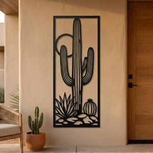 May include: Black metal wall art featuring a desert cactus scene. The artwork depicts a large saguaro cactus, a sun, and other desert plants. The piece is rectangular and mounted on a beige wall, suitable for indoor or outdoor decor.