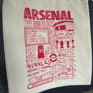 May include: A white canvas tote bag with a red Arsenal Football Club design. The design features the team's name, crest, and various statistics and achievements. The bag has two handles.