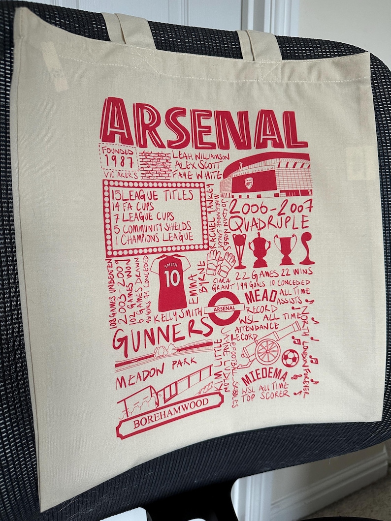 May include: A white canvas tote bag with a red Arsenal Football Club design. The design features the team's name, crest, and various statistics and achievements. The bag has two handles.