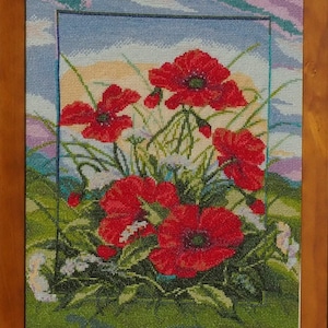 May include: A framed cross-stitch embroidery depicting a field of red poppies against a blue sky with white clouds. The flowers are in full bloom and the stems are green. The background is a field of green grass.