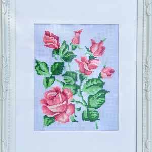 May include: A framed cross-stitch embroidery featuring a bouquet of pink roses with green leaves on a white background.