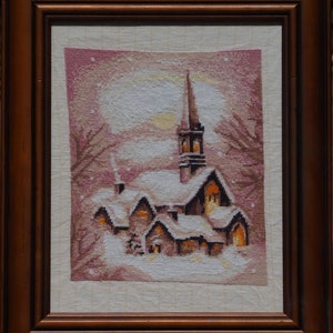 May include: A framed cross-stitch artwork depicting a snowy village scene with a church steeple and several houses. The houses have glowing windows and are covered in snow. The background is a soft pink with white snowflakes.