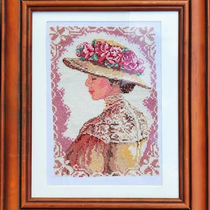 May include: A framed cross-stitch embroidery depicting a woman wearing a large straw hat with pink roses. The woman is wearing a white lace blouse and a brown dress. The embroidery is framed in a brown wooden frame.
