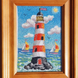 May include: Cross-stitch artwork of a lighthouse with red and white stripes, set against a blue sky with clouds, birds, and a yellow sun. Two sailboats are on the water. The artwork is framed in a wooden frame.
