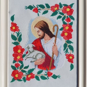May include: A framed cross-stitch embroidery depicting Jesus holding a lamb. The image is surrounded by a border of red flowers and green leaves.