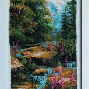 May include: Cross-stitch landscape art framed in white. The scene depicts a mountain range, a flowing stream, and lush greenery. Pink wildflowers add a pop of color to the detailed needlework.