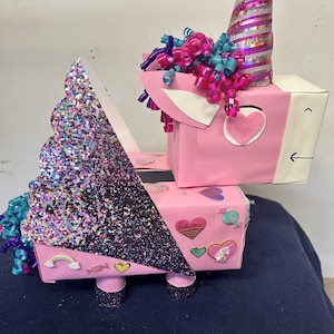 May include: A whimsical unicorn-shaped gift box, primarily pink, adorned with glitter, ribbons, and heart-shaped embellishments. The unicorn features a glittery horn and a mane of curly ribbons in shades of pink and blue. The box is decorated with stickers.