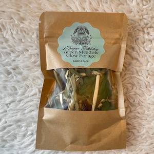 Op de afbeelding: Een bruine papieren zak met een doorzichtig venster met groen voer. De zak heeft een lichtblauw en wit etiket met de tekst "Morgan Rabbitry Green Meadow Glow Forage SAMPLE PACK" en een tekening van een konijn.