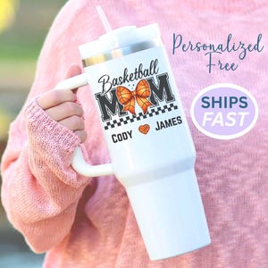 May include: White tumbler with a handle, featuring the text "Basketball MOM" with a basketball bow graphic. The names "Cody" and "James" are printed below. A straw is visible. The tumbler is held by a person wearing a pink sweater.