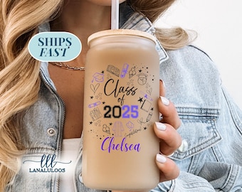 May include: A clear glass tumbler with a wooden lid and straw, featuring a graduation-themed design. The design includes the text "Class of 2025" and the name "Chelsea" in purple, along with graduation-related illustrations. The tumbler is held by a person.