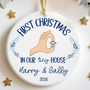 May include: A white ceramic Christmas ornament with the text "First Christmas in our tiny house" and the names "Harry & Sally" and the year "2026". The ornament features a hand holding a tiny house illustration and is decorated with blue floral accents and gold star ornaments.