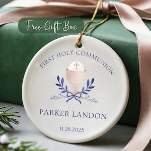 May include: A white ceramic First Holy Communion ornament with a gold string, featuring a chalice and cross design. The ornament includes the text "First Holy Communion", a name, and a date. It is presented with a green gift box and a pink ribbon.