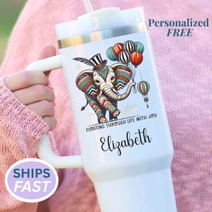 May include: White insulated tumbler with a handle, featuring a colorful elephant illustration with balloons. The text "FLOATING THROUGH LIFE WITH JOY" and a name are printed on the tumbler. The words "Personalized FREE" are also visible.