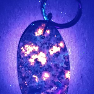 May include: Oval-shaped keychain with a dark background and a speckled pattern. The pattern has orange and yellow glowing spots, with a metal ring at the top. The keychain is likely made of a resin or stone material.