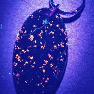 May include: An oval-shaped pendant with a dark blue background, speckled with orange and yellow flecks. The pendant is attached to a silver-colored key ring. The image is lit with a blue light.