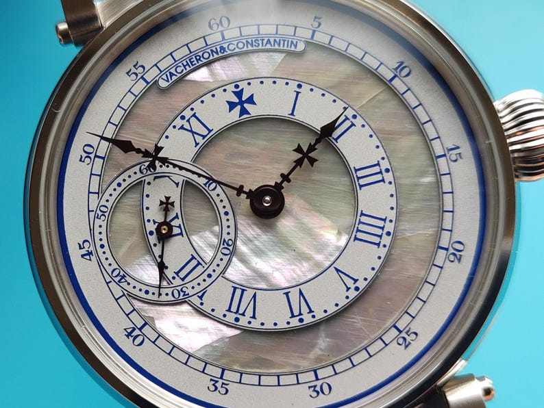 Pode incluir: Close-up de um rel&oacute;gio de luxo com caixa prateada e mostrador em madrep&eacute;rola. O rel&oacute;gio apresenta algarismos romanos, detalhes azuis e um submostrador. A marca "VACHERON & CONSTANTIN" &eacute; vis&iacute;vel.