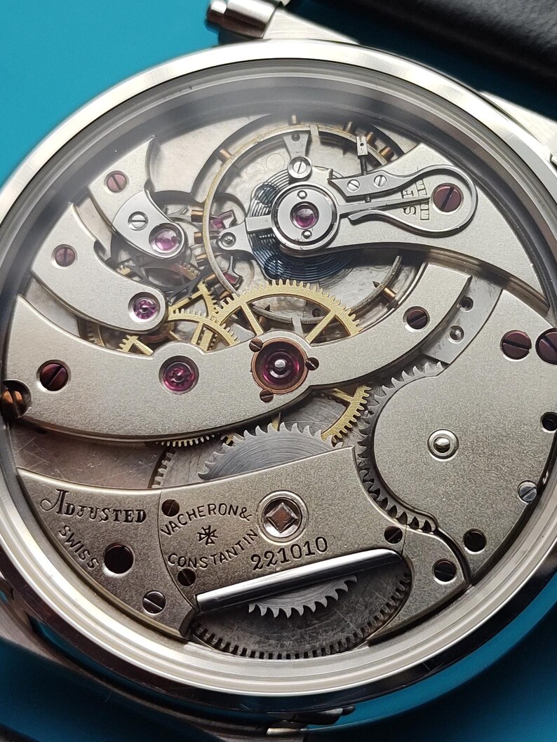 Pode incluir: Close-up do mecanismo de um rel&oacute;gio de pulso prateado com engrenagens intrincadas, joias e o texto "Adjusted Swiss Vacheron & Constantin 221010". Os componentes internos do rel&oacute;gio s&atilde;o vis&iacute;veis atrav&eacute;s de um cristal transparente.