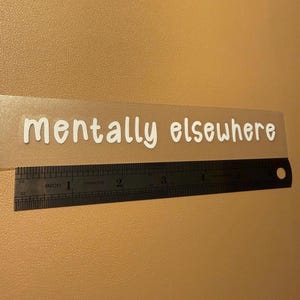 Może przedstawiać: Przezroczysta naklejka z napisem "mentally elsewhere" w białej, bezszeryfowej czcionce. Czarna linijka z pomiarami w calach znajduje się poniżej, mierząc około 12,7 cm. Tło jest w jednolitym kolorze beżowym.