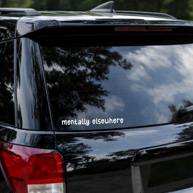 Może przedstawiać: Tylna szyba czarnego SUV-a z białym napisem "mentally elsewhere". W oknie odbija się zachmurzone niebo i gałęzie drzew. Tylne światło jest czerwone.