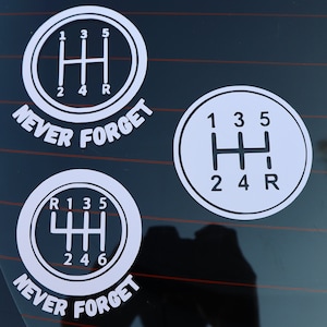 Puede incluir: Tres pegatinas de vinilo blanco con un patrón de cambio de marchas y el texto "NEVER FORGET" en la parte inferior. El patrón de cambio de marchas muestra los números 1, 2, 3, 4, 5 y R.