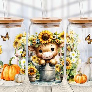 May include: Three clear glass tumblers with bamboo lids and glass straws. The designs feature a fluffy brown calf wearing a sunflower crown, orange and white pumpkins, sunflowers, daisies, and butterflies in a colorful, detailed illustration.