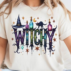 Puede incluir: Camiseta color crema con la palabra "WITCHY" en una fuente decorativa, adornada con ilustraciones temáticas de brujas. El diseño incluye un sombrero de bruja, botellas de pociones, un gato negro, estrellas y una luna creciente.
