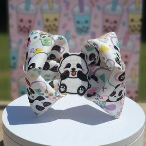 May include: A large hair bow made of white ribbon featuring a repeating pattern of pandas and colorful geometric shapes. A central patch depicts a cartoon panda with a pink tongue sticking out, black ears, and black limbs on a white background.