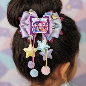 May include: A multi-layered hair bow featuring a central purple glitter patch with the text HUNTRIX and three cartoon characters. The bow includes iridescent lavender fabric, gold heart accents, and dangling ribbons with metallic stars and small pom-poms.