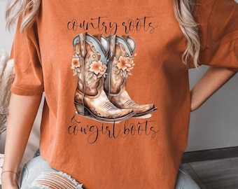May include: Orange t-shirt featuring a graphic of brown cowboy boots with floral accents. The text "country roots & cowgirl boots" is written above and below the boots in a script font. The shirt has short sleeves.