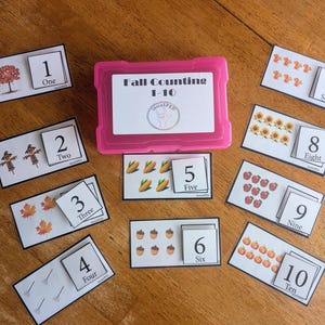 May include: A pink plastic storage box labeled Fall Counting 1-10 with a circular logo, surrounded by ten rectangular white cards. Each card features a number from one to ten, the corresponding written word, and illustrations of fall items.