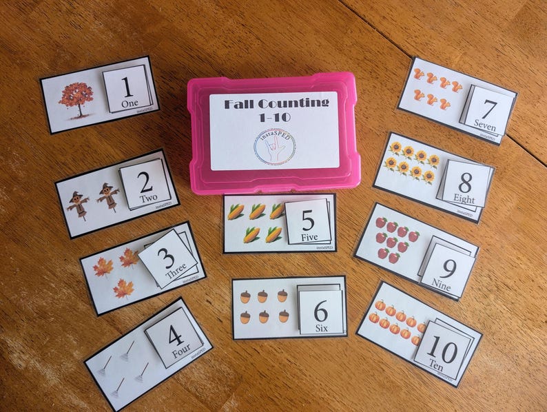 May include: A pink plastic storage box labelled Fall Counting 1-10 with a circular logo, surrounded by ten rectangular white cards. Each card features a number from one to ten, the corresponding written word, and illustrations of autumn items.