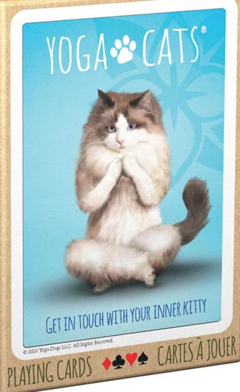 May include: A deck of playing cards featuring a white cat with brown and gray markings sitting in a yoga pose. The card has a blue background with a white paw print and the text "Yoga Cats" in white. The text "Get in touch with your inner kitty" is printed below the cat. The bottom of the card has the text "Playing Cards" and "Cartes &agrave; Jouer" in brown.